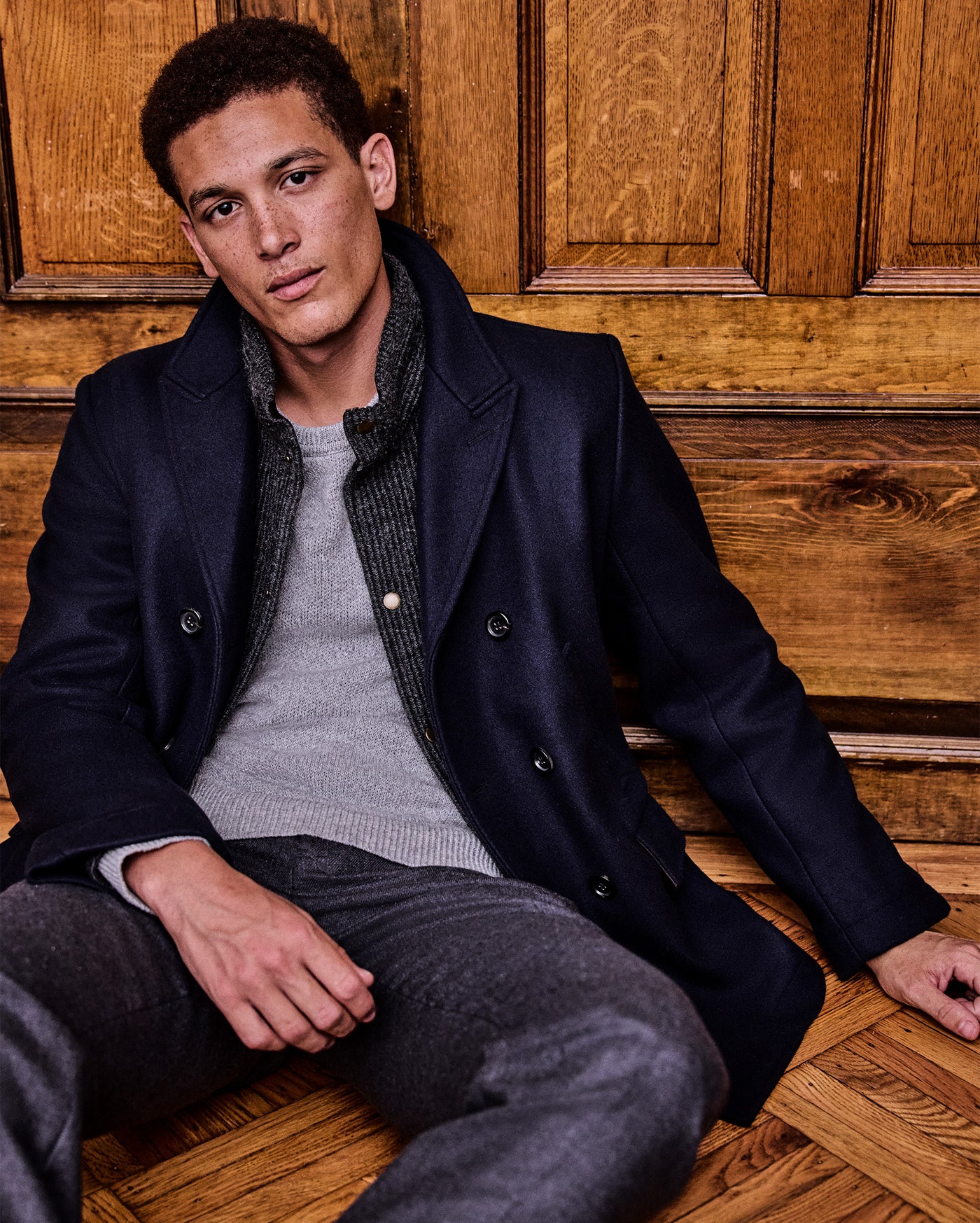 Man wearing a dark coat sitting on a wooden floor with wooden paneling.