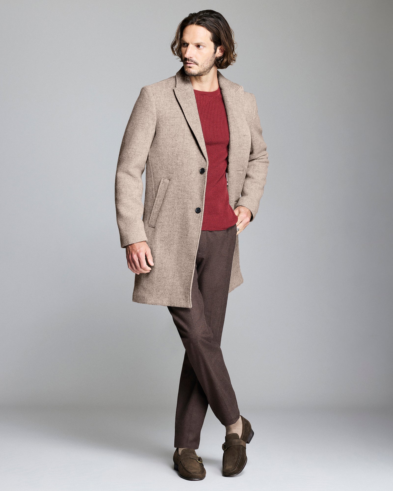 Man wearing a beige overcoat, red shirt, and brown pants on a gray background