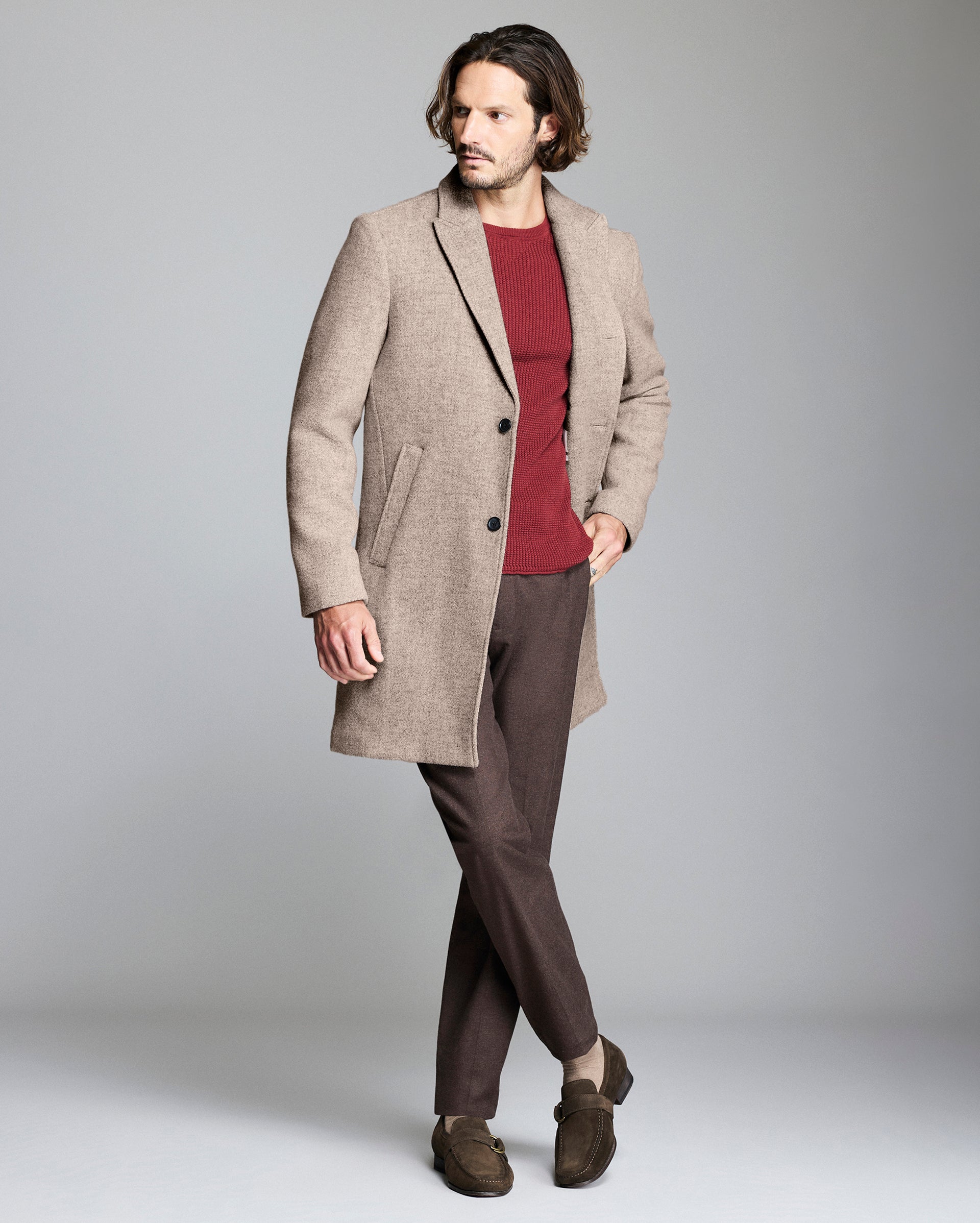 Man wearing a beige overcoat, red shirt, and brown pants on a gray background