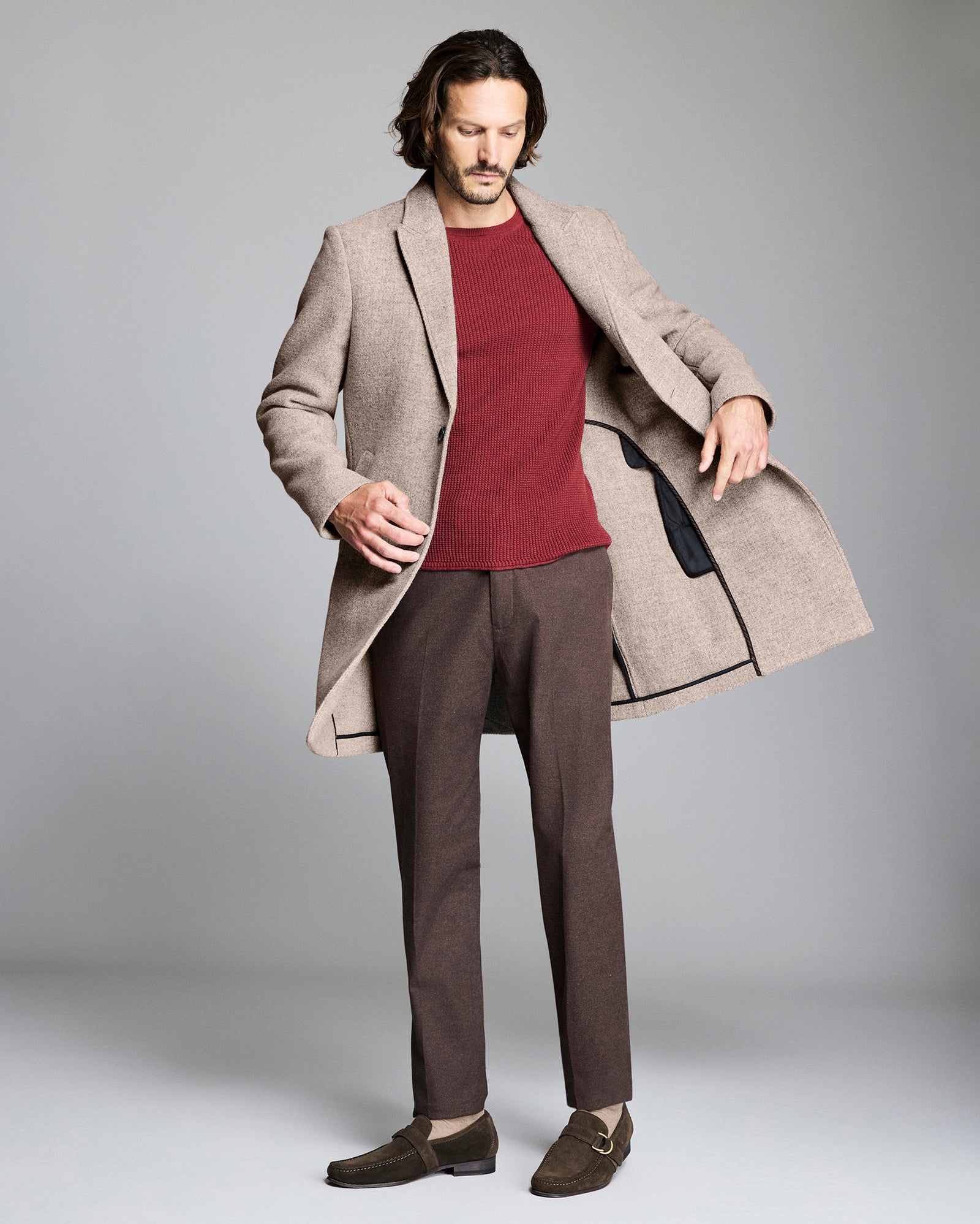 Man wearing a beige coat over a red sweater and brown pants on a gray background