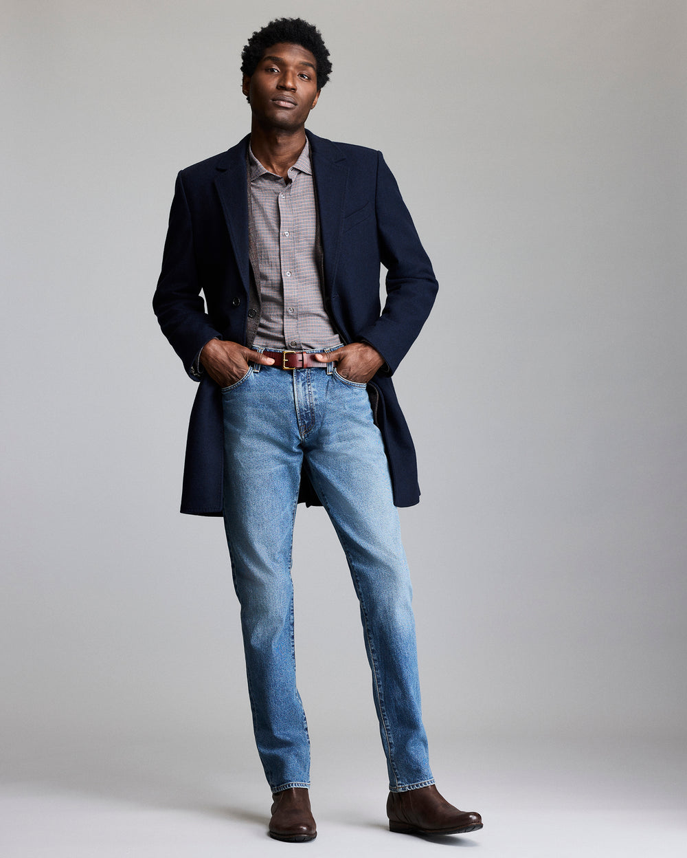 Man wearing a navy coat, light gray shirt, blue jeans, and brown shoes on a plain background