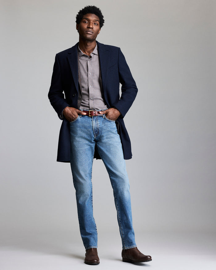 Man wearing a navy coat, light gray shirt, blue jeans, and brown shoes on a plain background