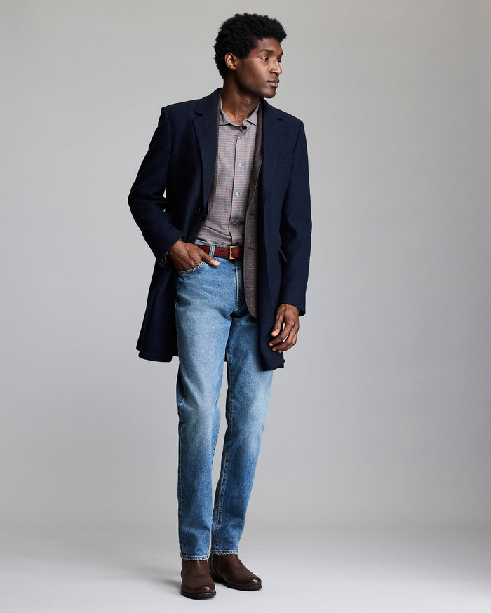Man wearing a navy coat, light shirt, blue jeans, and brown shoes on a plain background