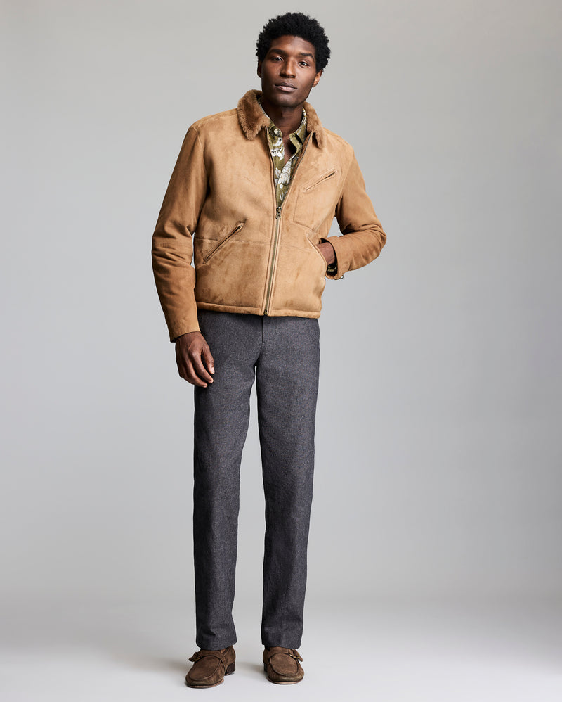 Man wearing a tan suede jacket with fur collar and gray pants on a plain background Alternate