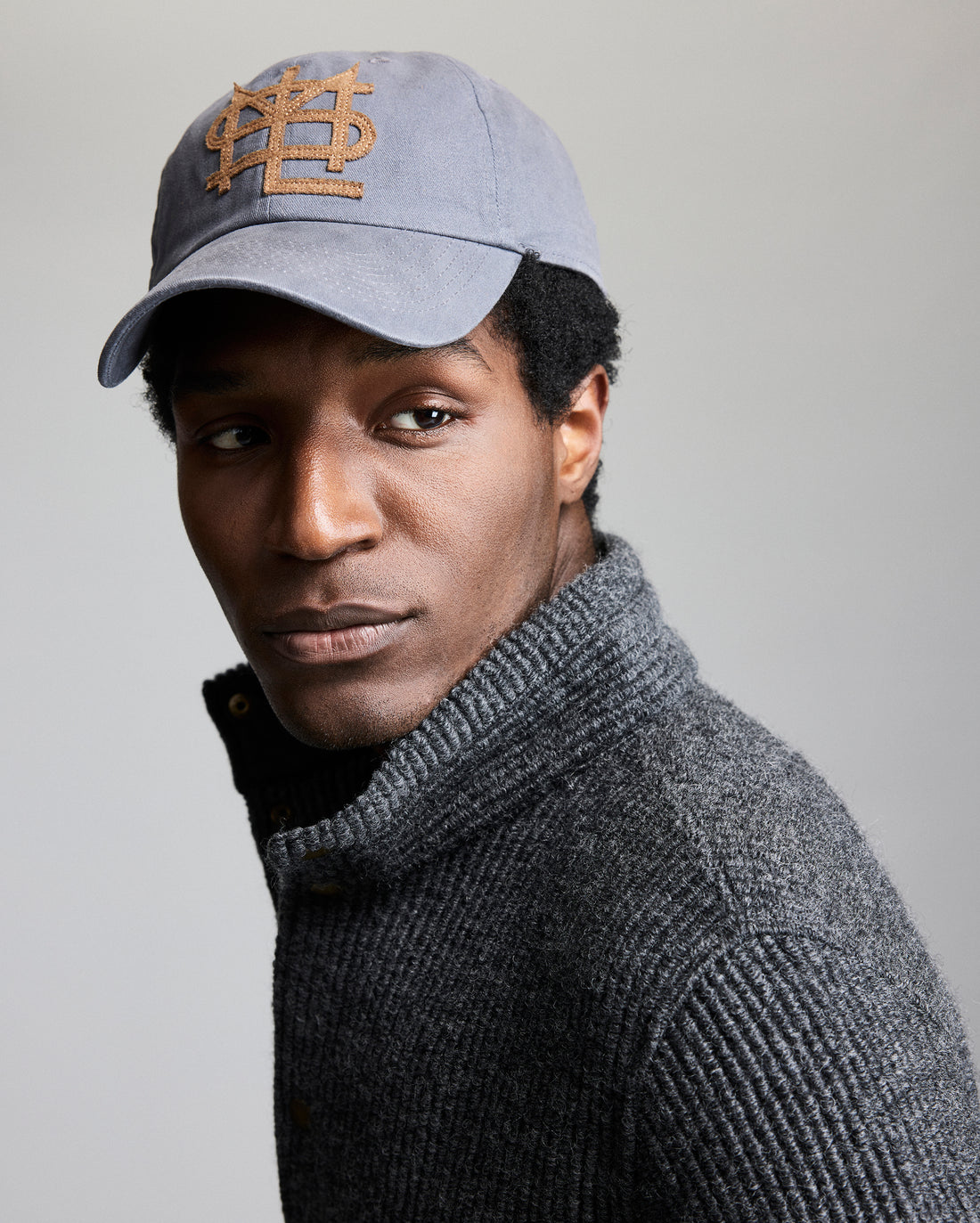 Man wearing a gray cap with a logo and a gray sweater on a plain background
