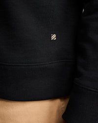 Close-up of a person wearing a black sweatshirt and tan pants. The sweatshirt features a small rectangular tag with diagonal stripes near the seam above the waistband. The persons arm and part of their side are visible.