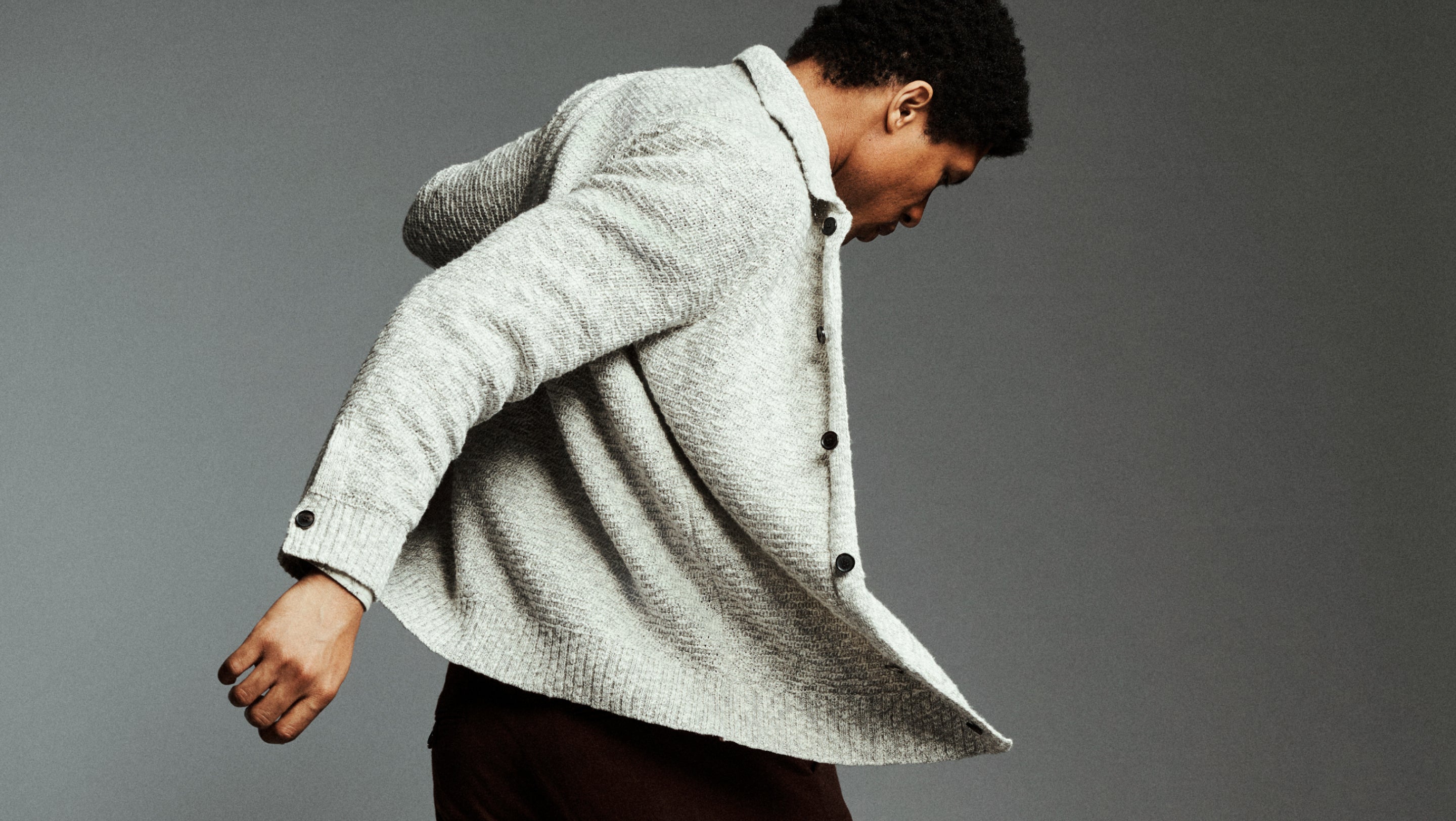 A man wearing a light gray textured knit cardigan sweater and dark brown trousers, captured in a dynamic pose against a solid gray studio background.