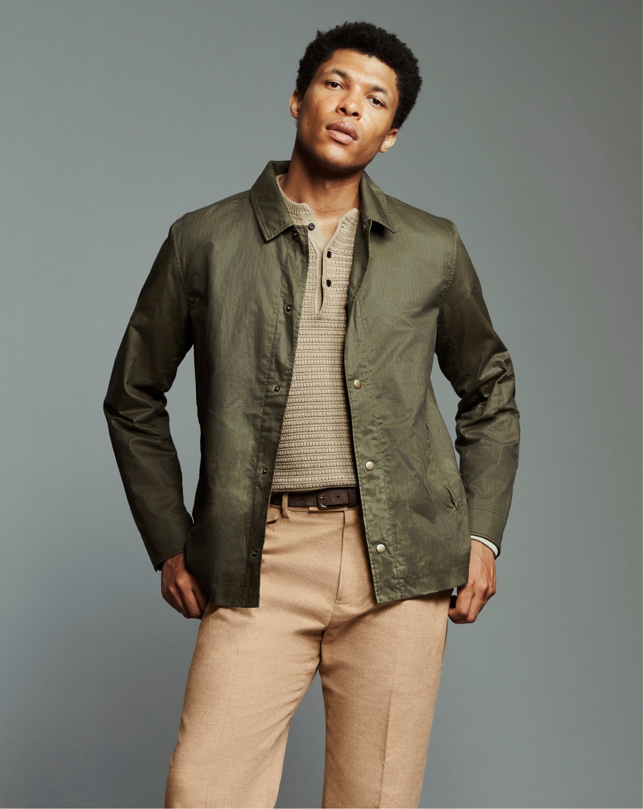 A man modeling an olive green lightweight button-up jacket over a beige knit polo shirt and tan trousers, photographed against a solid gray studio background.