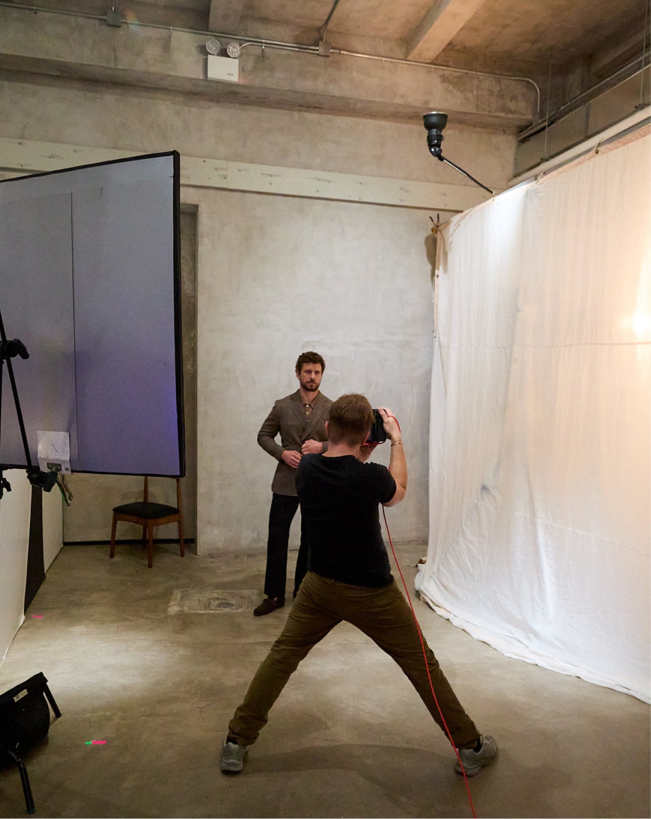 A man in a black t-shirt and olive pants points a camera at a male model. The model wears a gray blazer and stands in the background of a studio setup with a large white drape and screen.