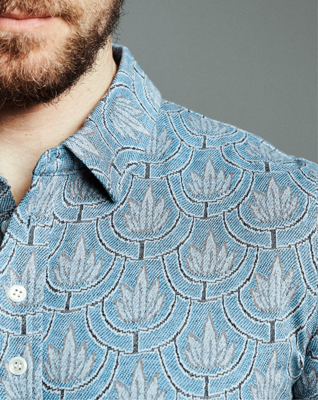 Close-up of a man wearing a light blue polo with a repeating gray and white leaf pattern, showing the collar, fabric texture, and part of his beard against a neutral gray background.