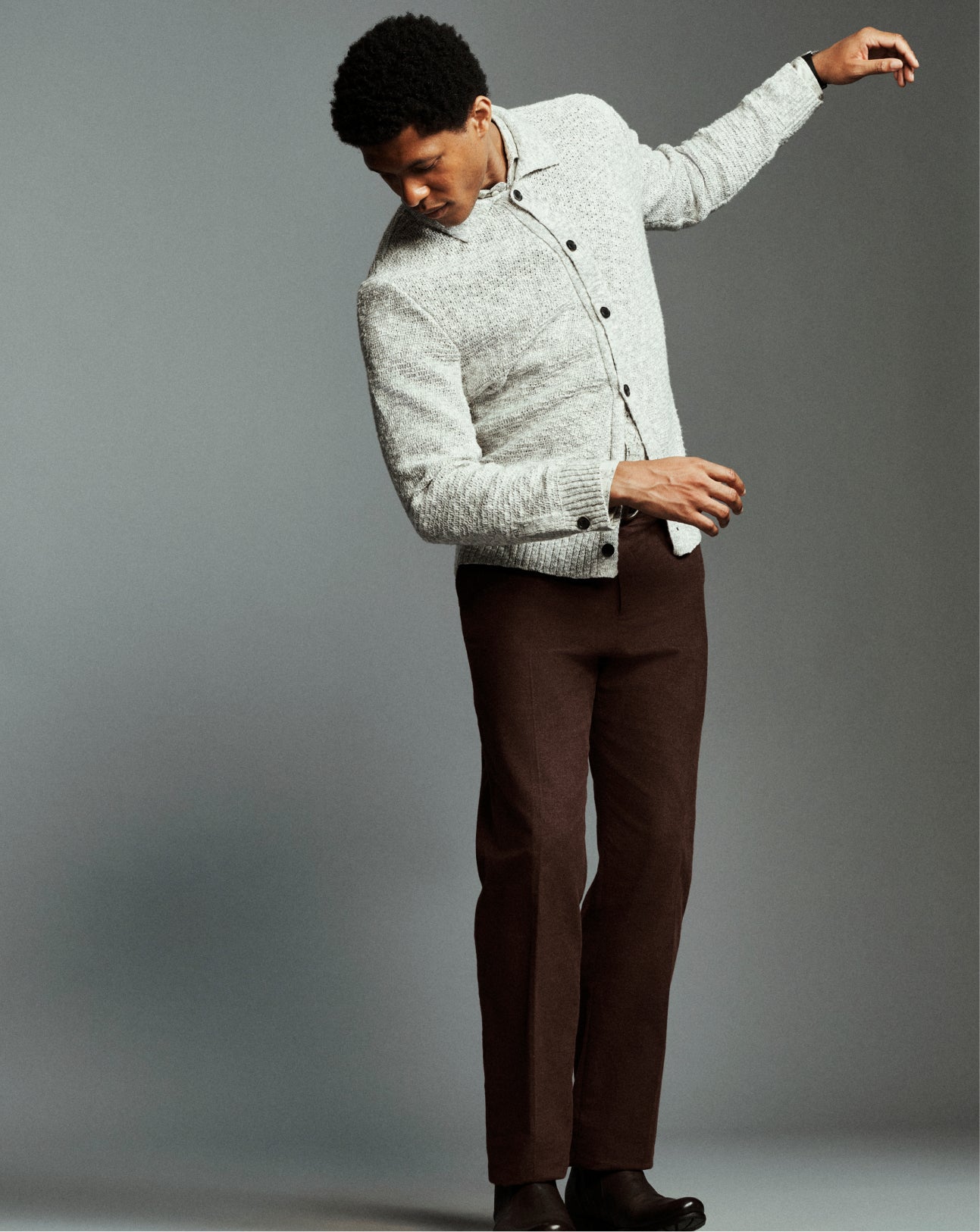 A man modeling a light gray textured knit button-up shirt jacket with dark buttons, paired with dark brown trousers and black shoes, against a solid gray studio background.