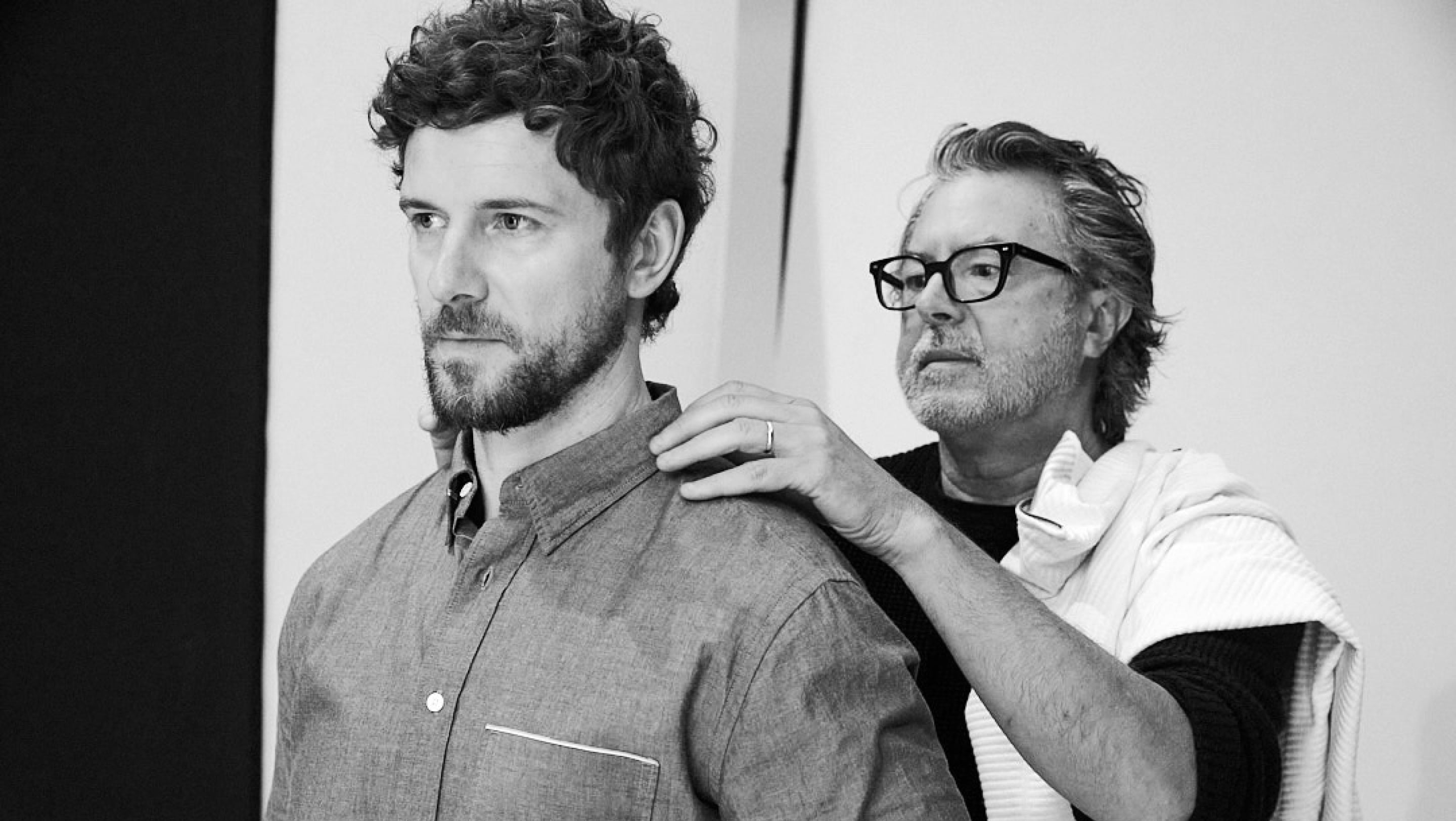 A black and white photograph of two men, one with curly hair and a beard wearing a button-down shirt, and the other, Billy Reid, is wearing glasses and a white shirt, with the man in the back placing his hand on the man in the front's shoulder to adjust his shirt.