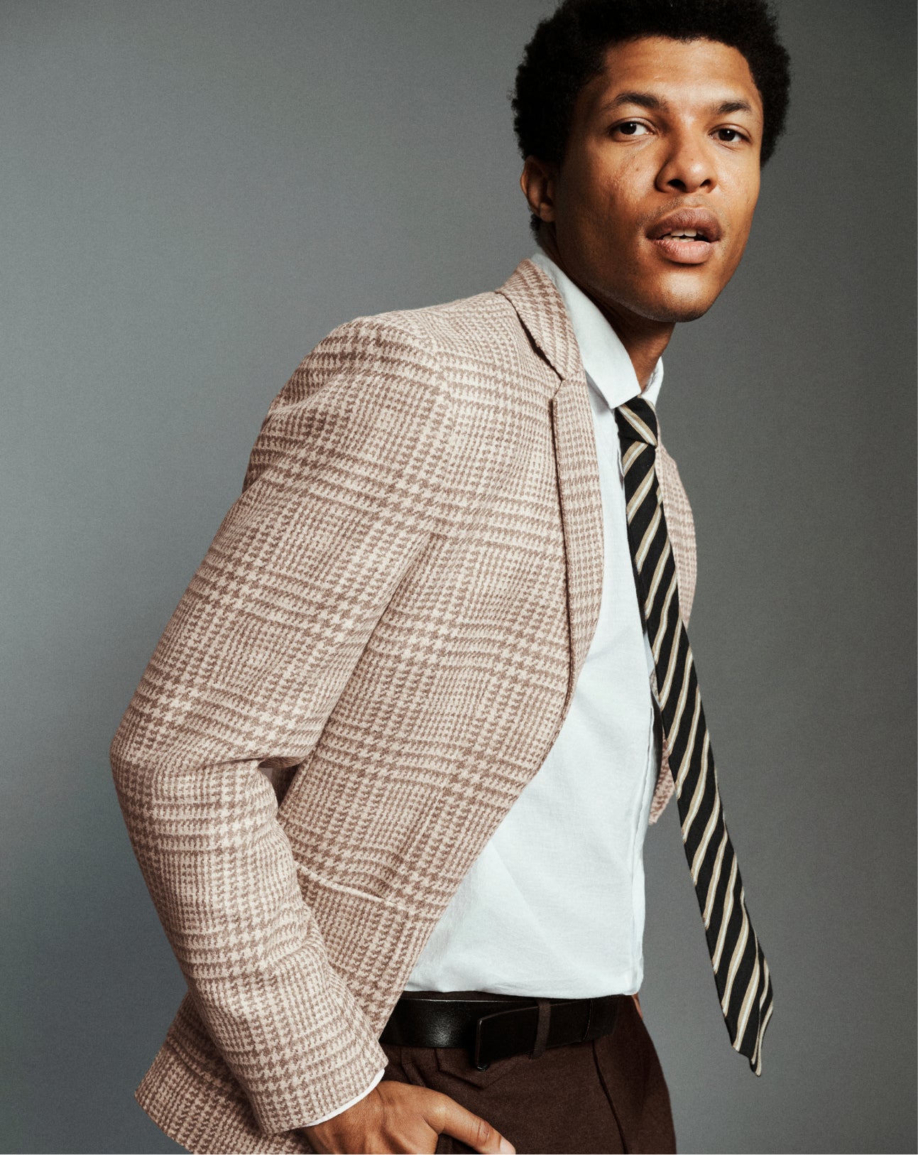 A man wearing a beige and brown checkered blazer, a white dress shirt, and a black and beige striped tie, posed against a solid gray studio background.
