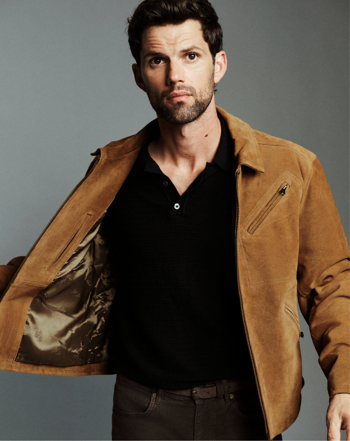 A man wearing a tan suede jacket over a black polo shirt and dark pants, posed against a plain gray studio background.