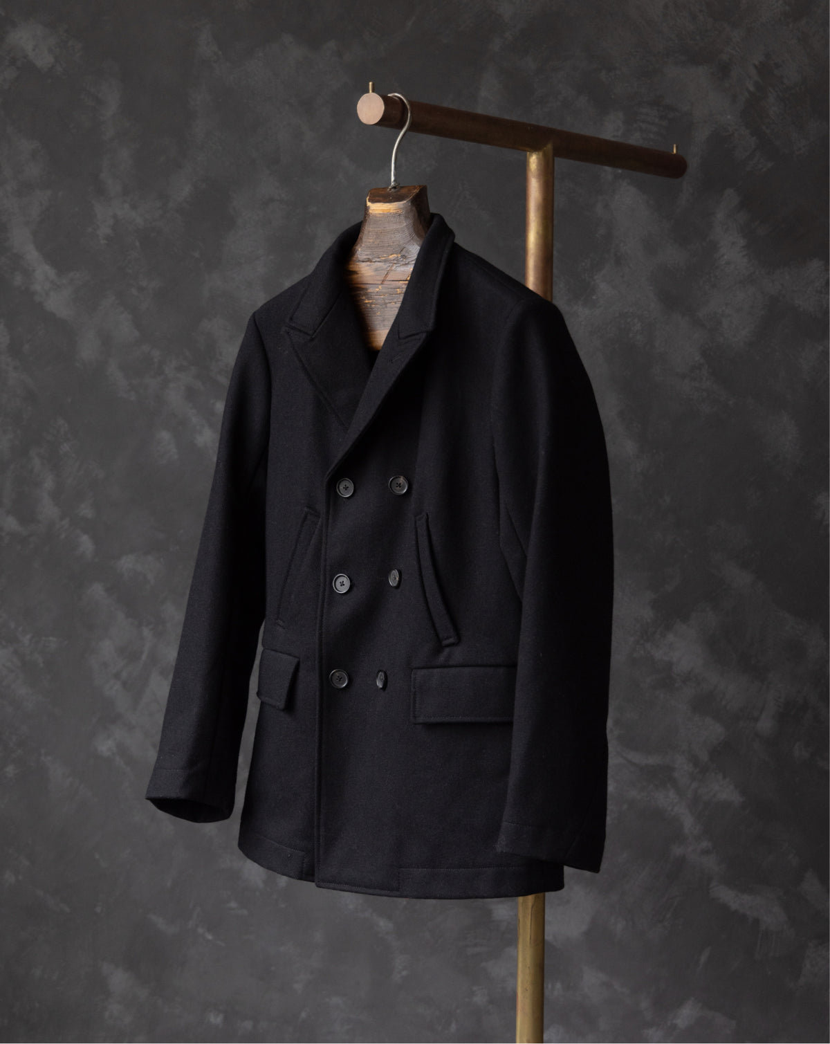 A black double-breasted wool-blend jacket with a notch lapel and dark buttons, hanging on a wooden hanger stand against a dark textured gray background.