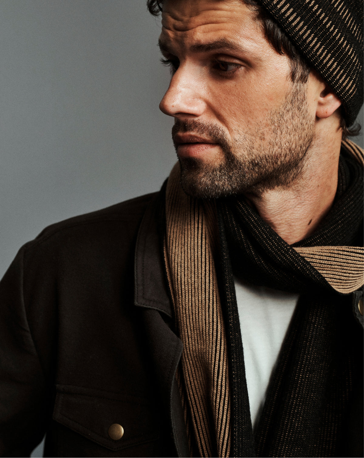 A close-up portrait of a man wearing a dark brown jacket, a beige and black striped knit beanie, and a dark brown striped knit scarf over a white shirt, set against a solid gray background.