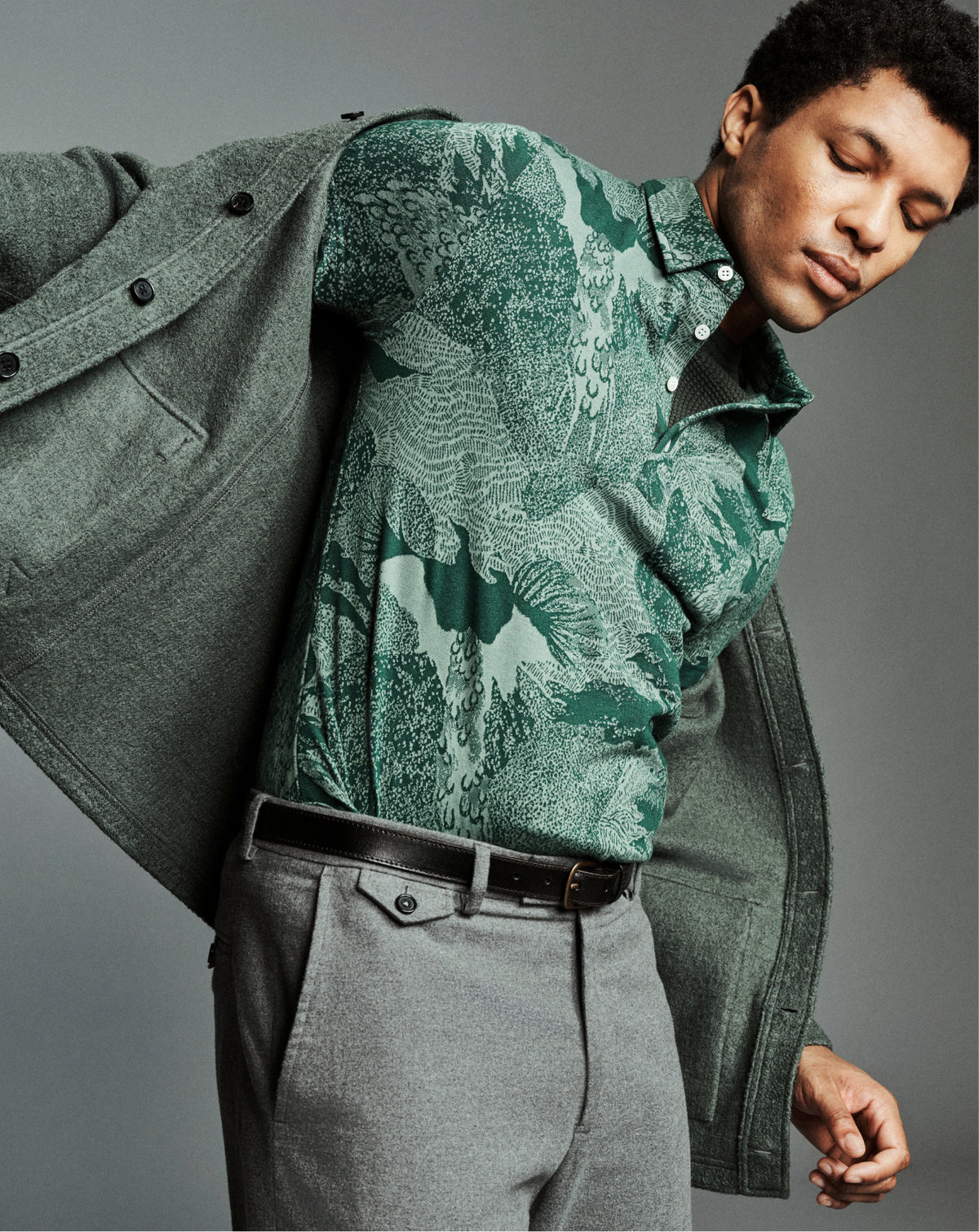 A man wearing a green patterned polo shirt, gray trousers, and an open olive green overshirt, posed against a neutral gray studio background.