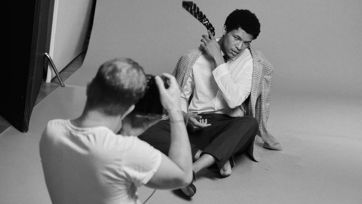 A black and white photograph showing a model sitting on the floor of a studio while a photographer takes a picture. The model is wearing a light-colored shirt and a checkered jacket, holding a guitar against his head.