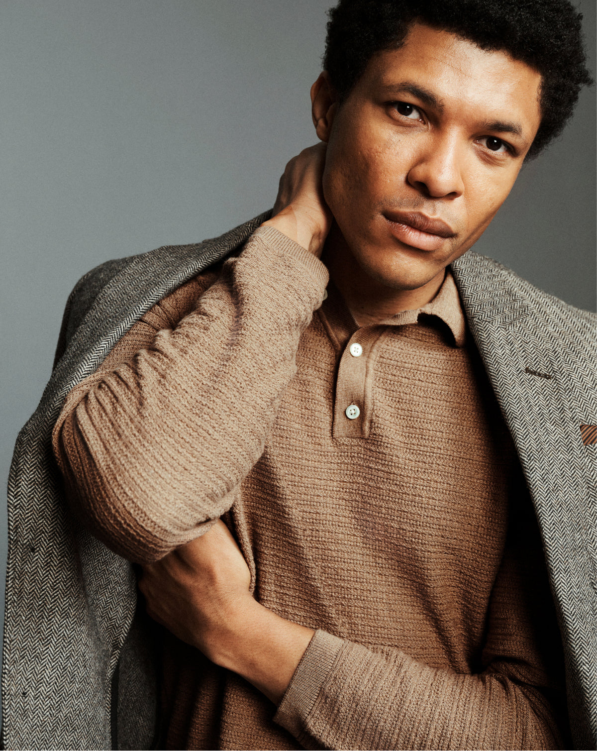A man wearing a brown ribbed knit polo shirt under a gray textured blazer, posing with his hand on his neck against a solid gray studio background.