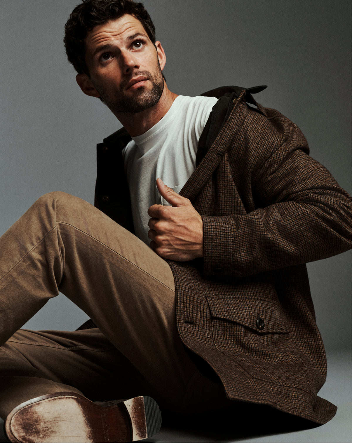 A man sitting on the floor wearing a brown textured coat over a white t-shirt and tan pants, looking off-camera against a dark gray studio background.
