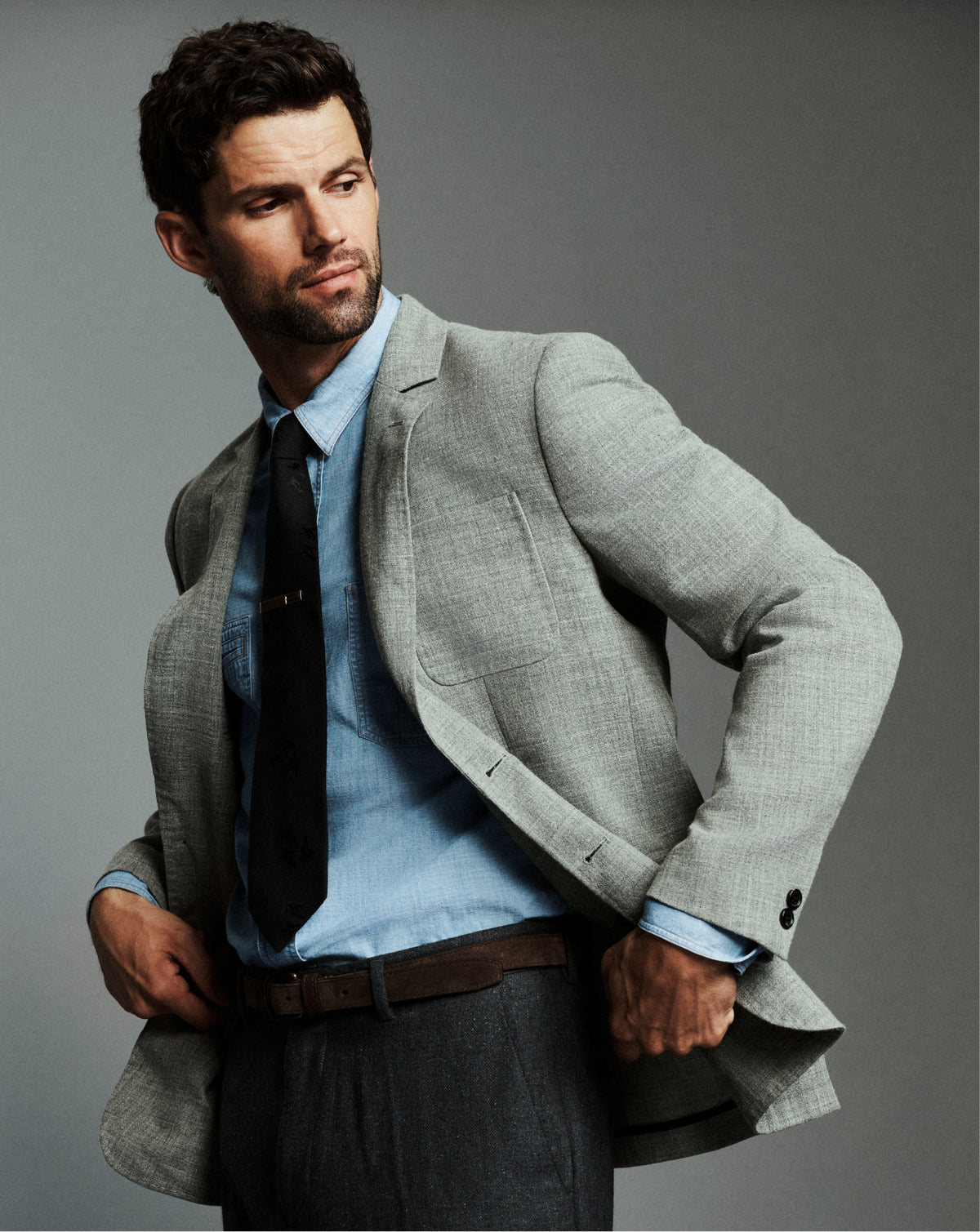 A man wearing a light gray textured blazer over a light blue dress shirt and black tie, paired with dark gray trousers and a brown belt, posed against a solid gray studio background.