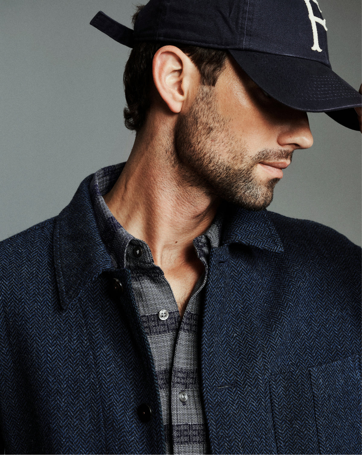 A close-up of a man wearing a navy blue herringbone patterned overshirt over a gray and white plaid button-down shirt, paired with a navy baseball cap featuring a white letter 'F'.