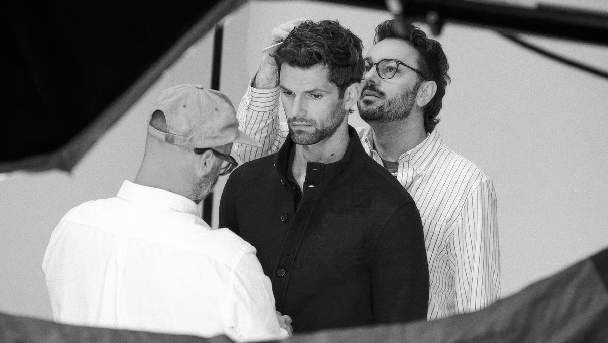 A black and white photograph showing three men in a studio setting, with the central figure wearing a dark button-up shirt and looking downward, flanked by two other men in light-colored shirts.