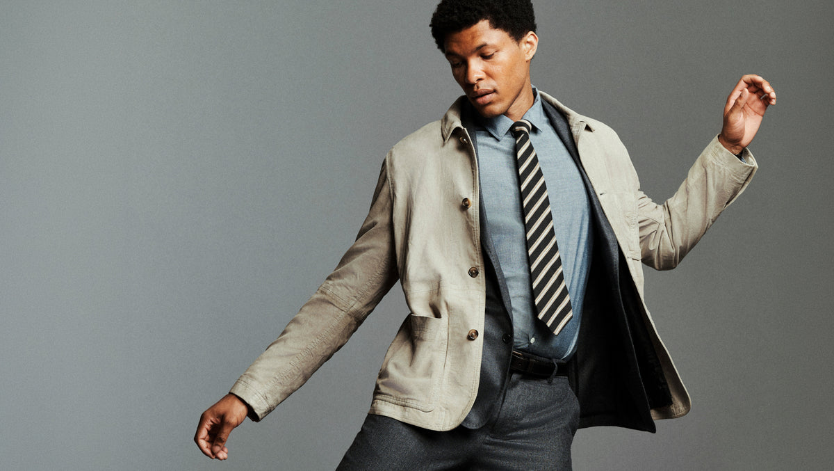 A man posing in a light beige coat, a blue shirt, a black and white striped tie, and dark gray trousers against a solid gray studio background.