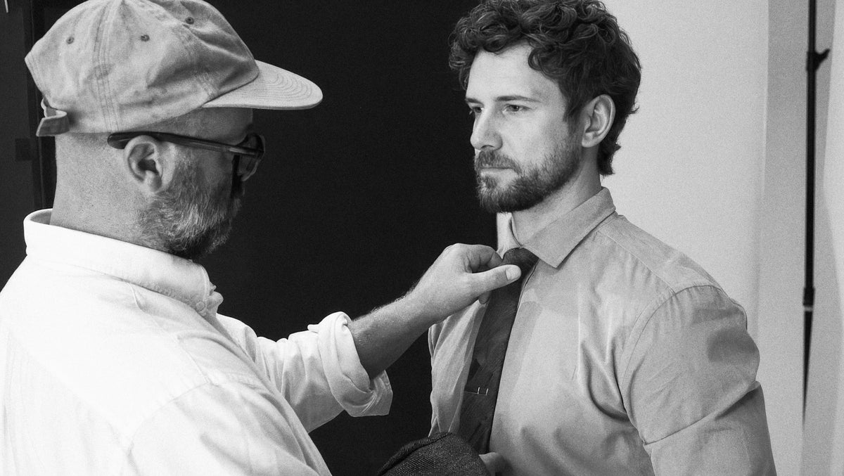A black and white photograph showing a man with a beard and cap adjusting the tie of another man wearing a button-down shirt.