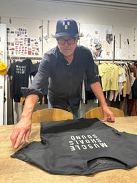 A man wearing a black baseball cap and glasses stands behind a table in a clothing studio, adjusting a black t-shirt laid flat. The t-shirt features the text 'MUSCLE SHOALS SOUND' printed in white on the front.