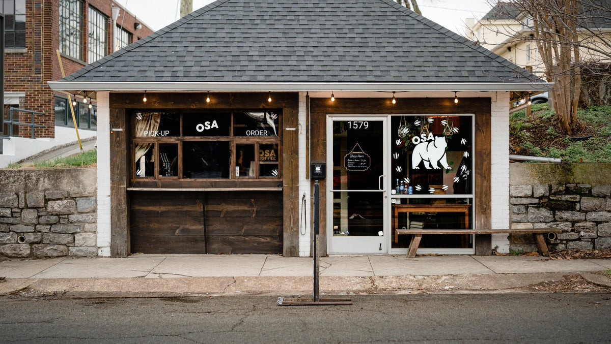 A small rustic coffee shop with a dark gray shingled roof, white trim, and dark wood paneling on a city street. The building has a window graphic of a bear with the restaurant name 'OSA' above it on the left of the glass door entrance. On the right is a walk-up pickup counter.