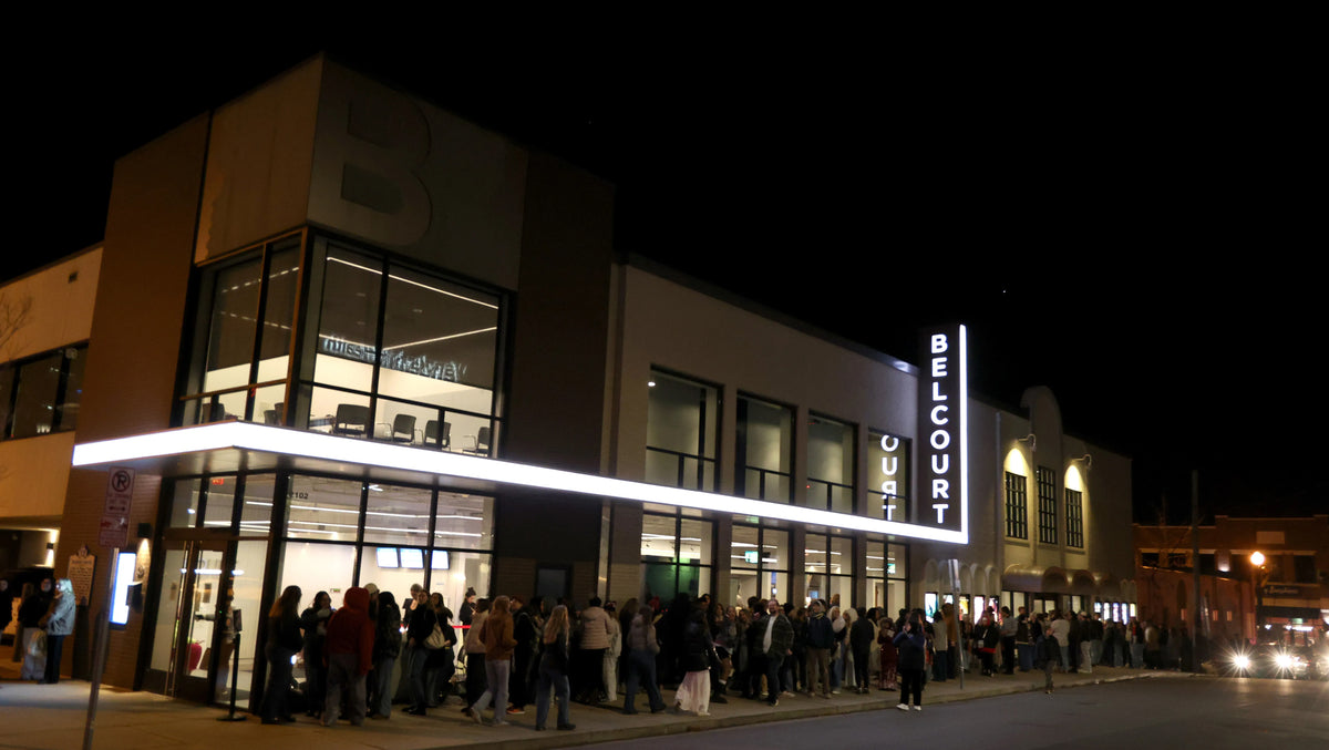 A nighttime street scene showing a modern building with large glass windows and a vertical illuminated sign reading 'BELCOURT'. A long line of people is visible along the sidewalk in front of the building.