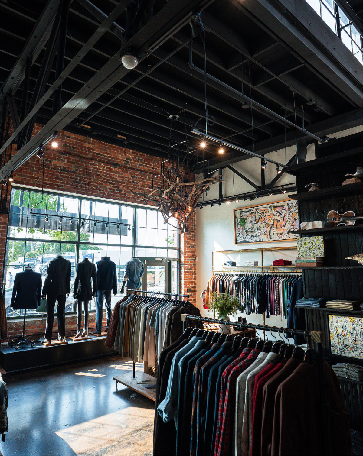 A clothing store interior with racks of shirts and suits, mannequins displaying dark formal wear, a brick wall, exposed ceiling beams, and large windows.