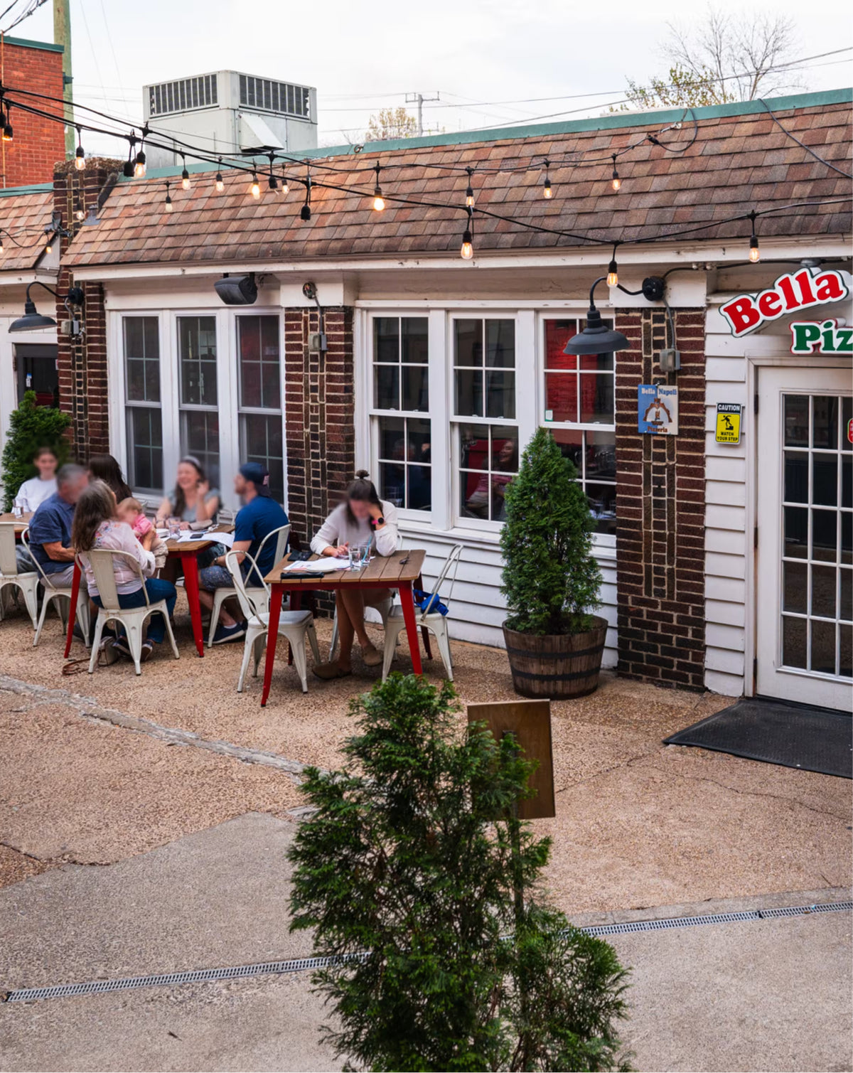 Outdoor dining area at Bella Pizzeria with several people seated at tables on a gravel patio in front of a brick and white siding restaurant building.