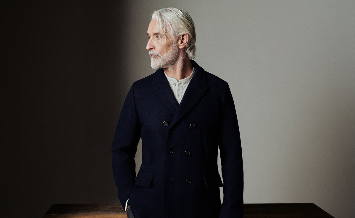 A man with white hair and a beard wearing a dark navy double-breasted coat over a light-colored shirt, standing in profile against a dark and light gray studio background.