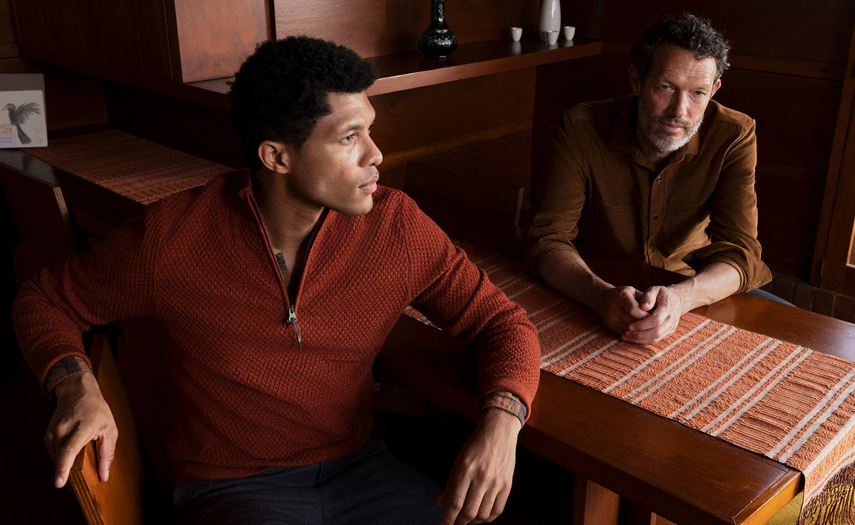 Two men seated at a wooden table in a warm-toned interior. The man in the foreground wears a rust-red textured half-zip sweater, while the man behind him wears a brown button-up shirt.