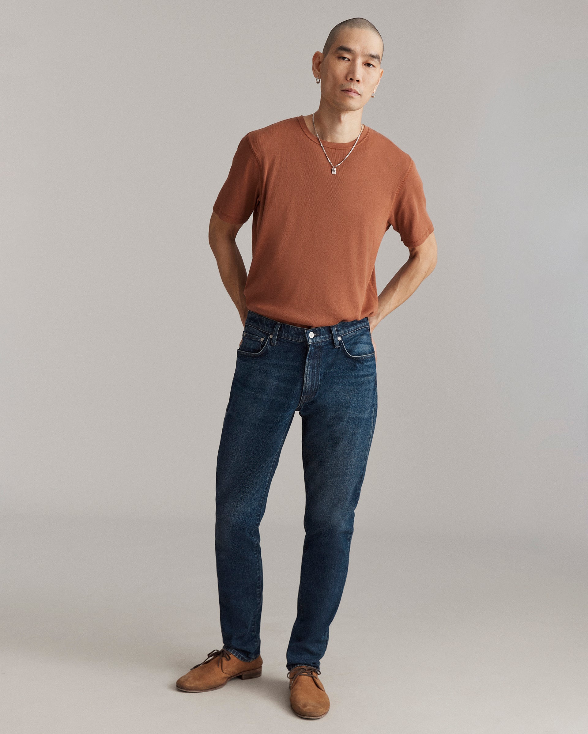 A person with short hair stands against a plain backdrop, wearing a rust-colored t-shirt, dark blue jeans, brown shoes, and a necklace, with hands behind their back and a neutral expression.