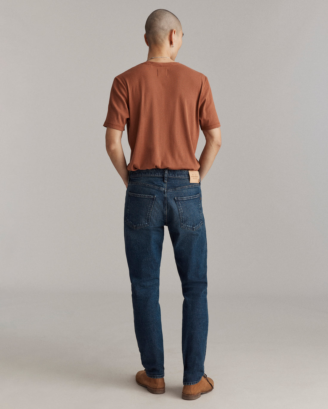 A person stands facing away from the camera, wearing a brown short-sleeve shirt, blue jeans, and brown shoes, against a plain light gray background.