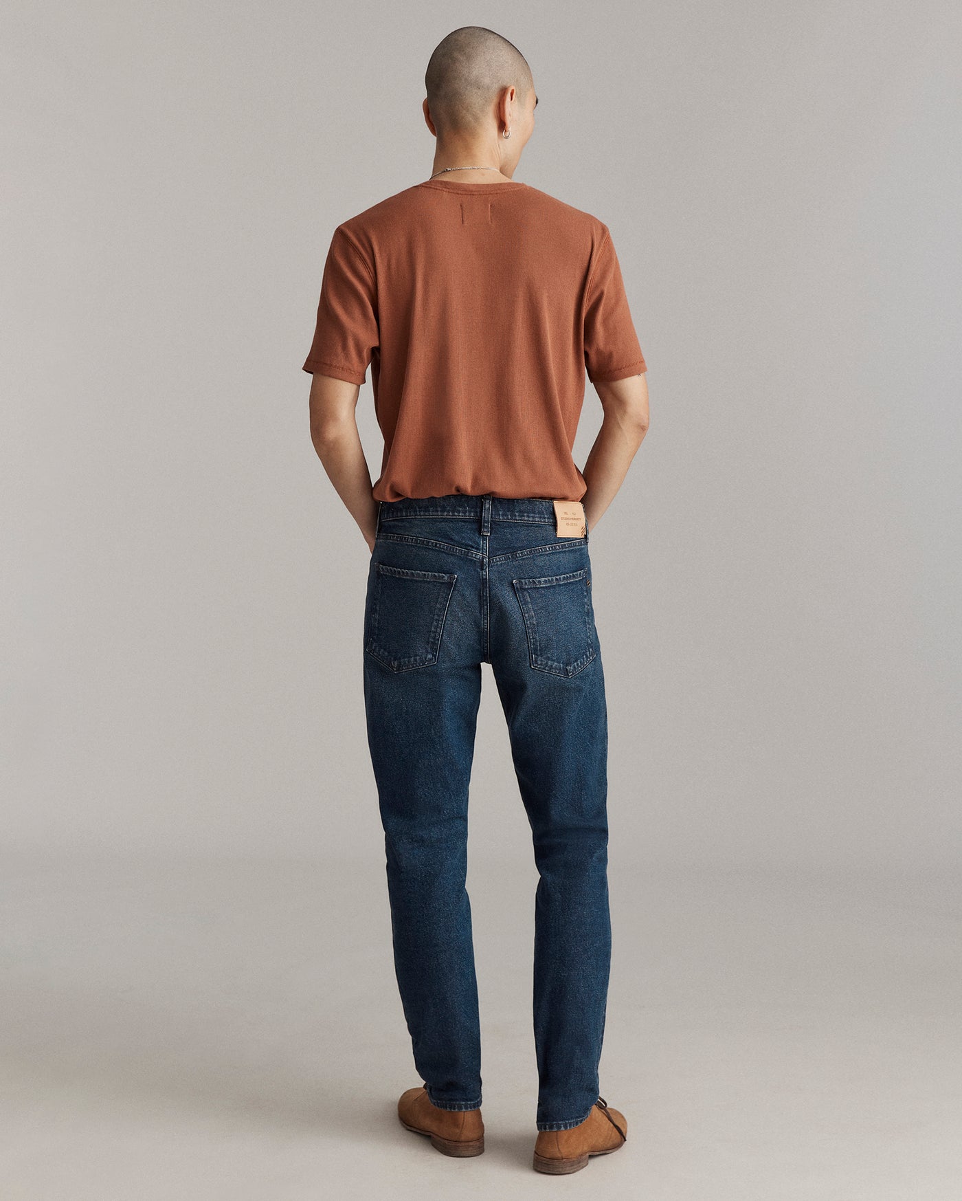 A person stands facing away from the camera, wearing a brown short-sleeve shirt, blue jeans, and brown shoes, against a plain light gray background.