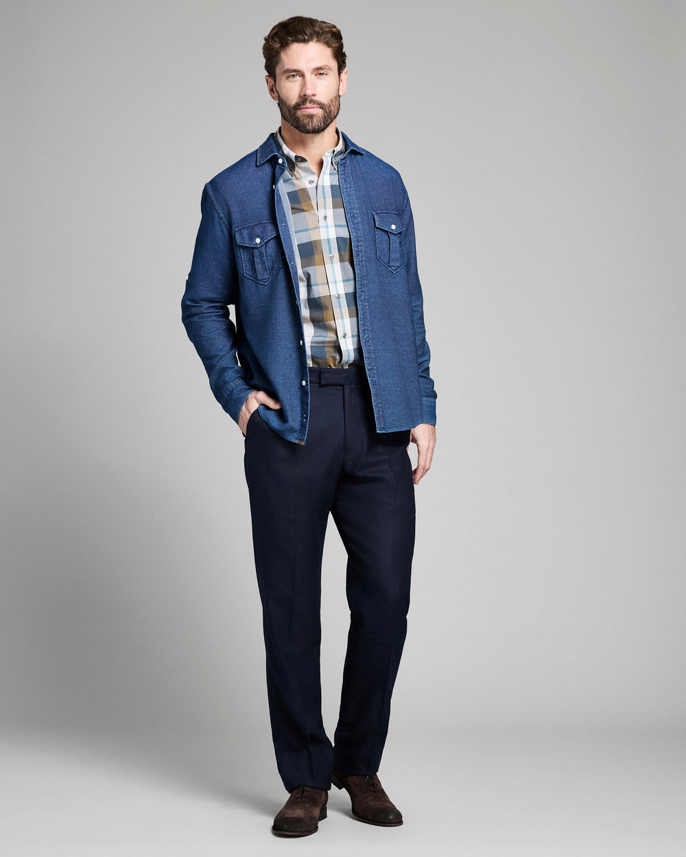 Man wearing a blue denim jacket over a plaid shirt, paired with dark pants and shoes on a gray background
