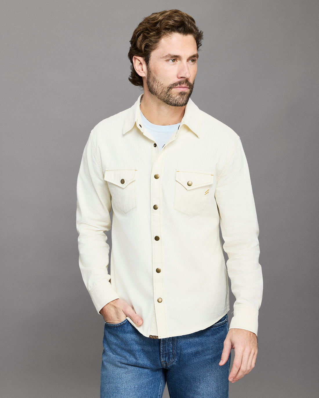 Man wearing a cream-colored denim jacket and blue jeans against a gray background