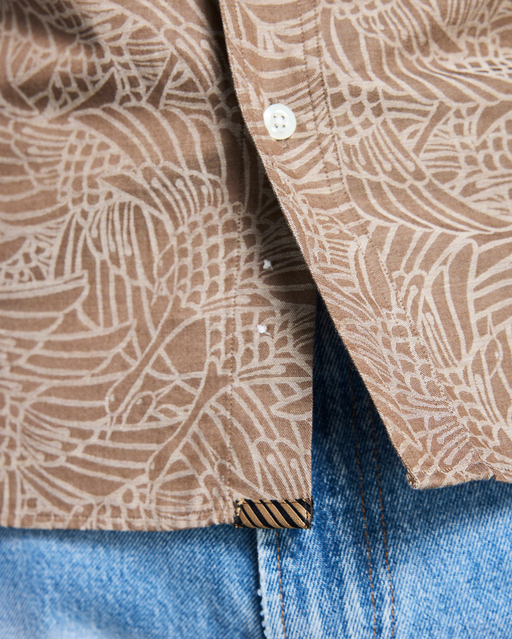 Close-up of a brown patterned shirt collar with a button, worn over blue jeans.