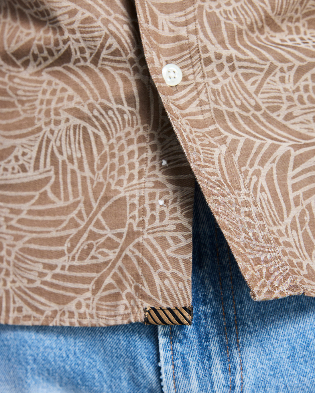 Close-up of a brown patterned shirt collar with a button, worn over blue jeans.