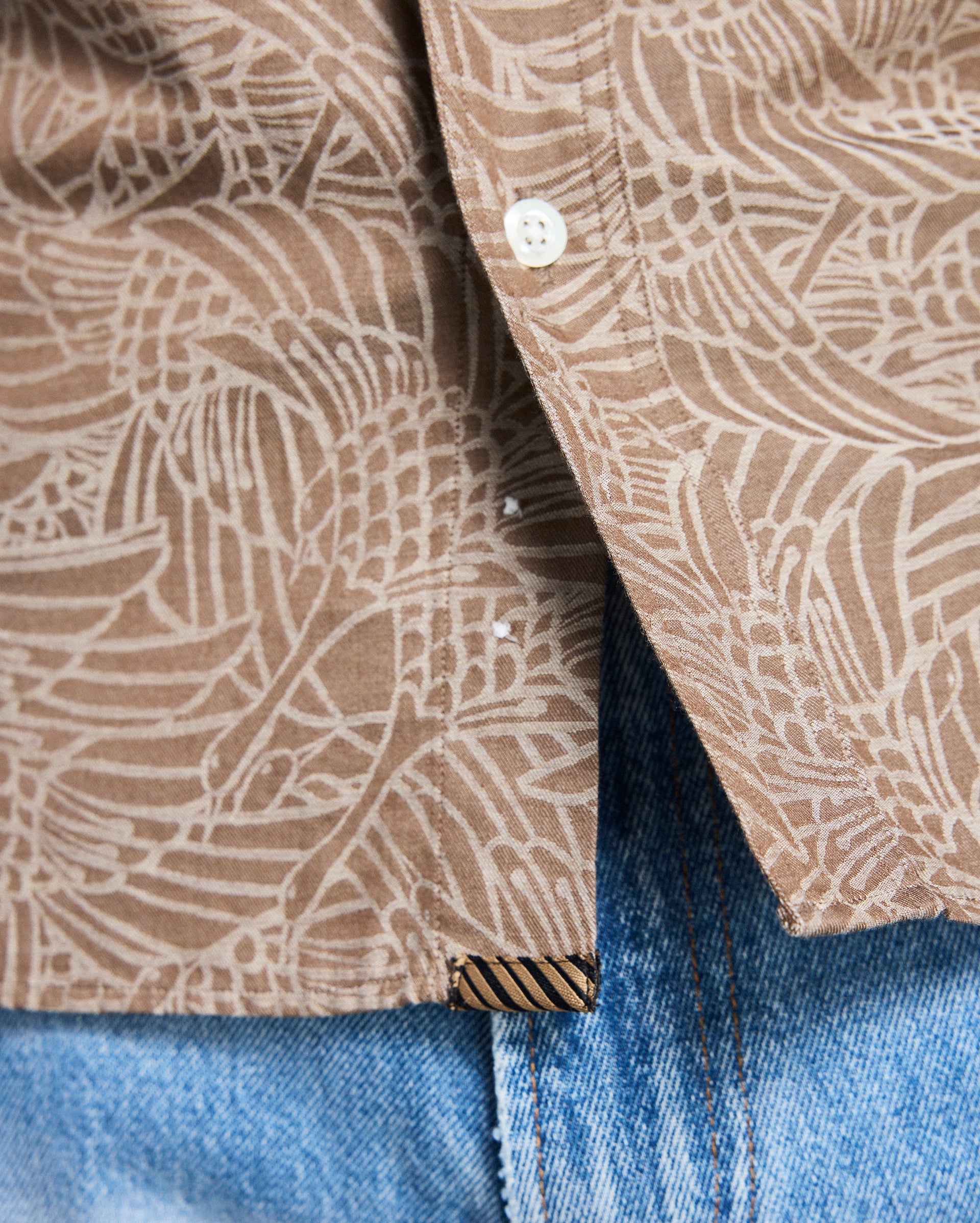 Close-up of a brown patterned shirt collar with a button, worn over blue jeans.
