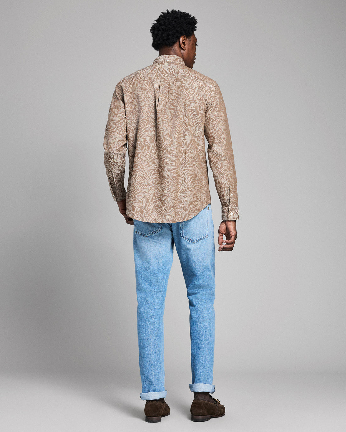 Man wearing a beige patterned shirt and light blue jeans on a gray background