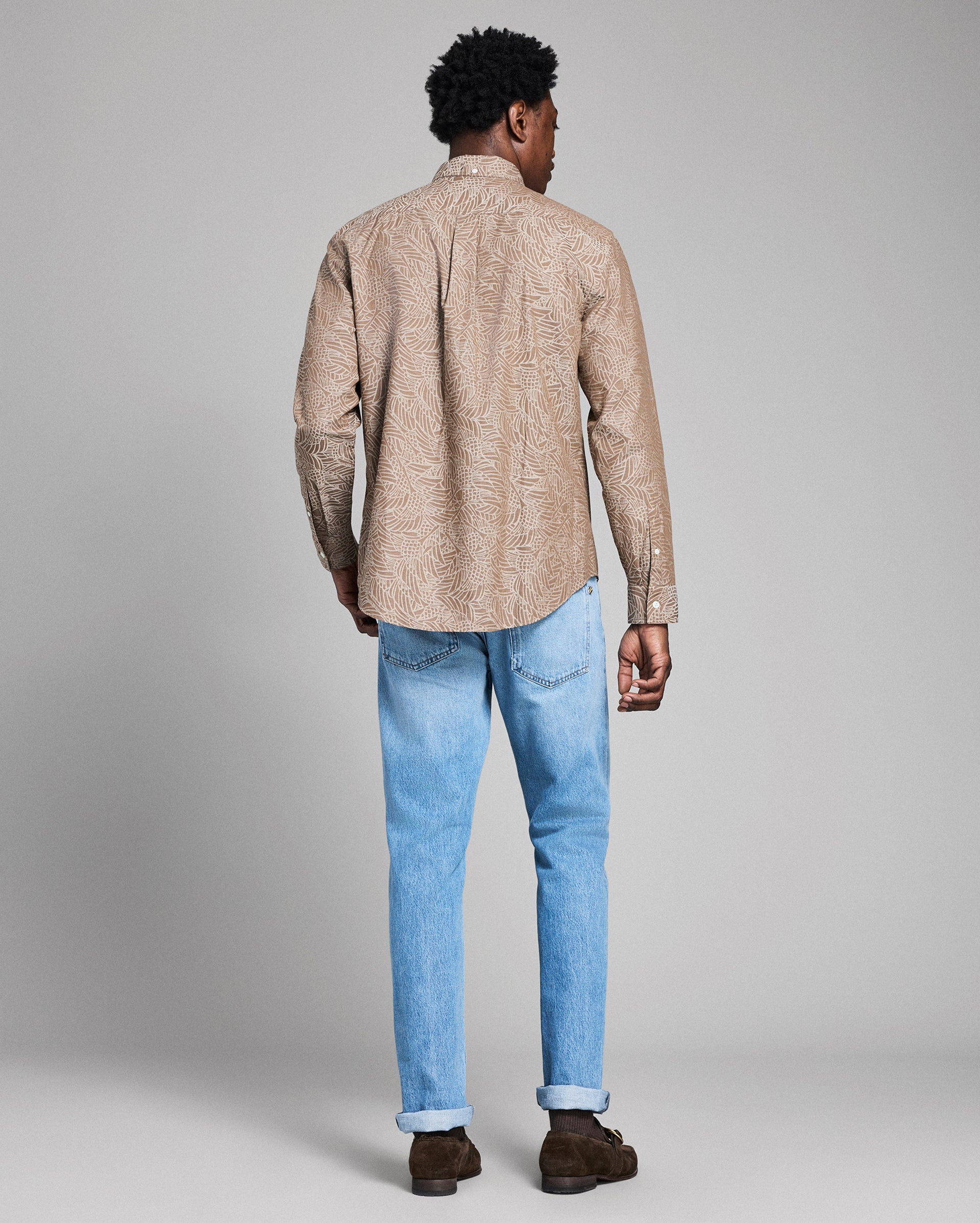 Man wearing a beige patterned shirt and light blue jeans on a gray background