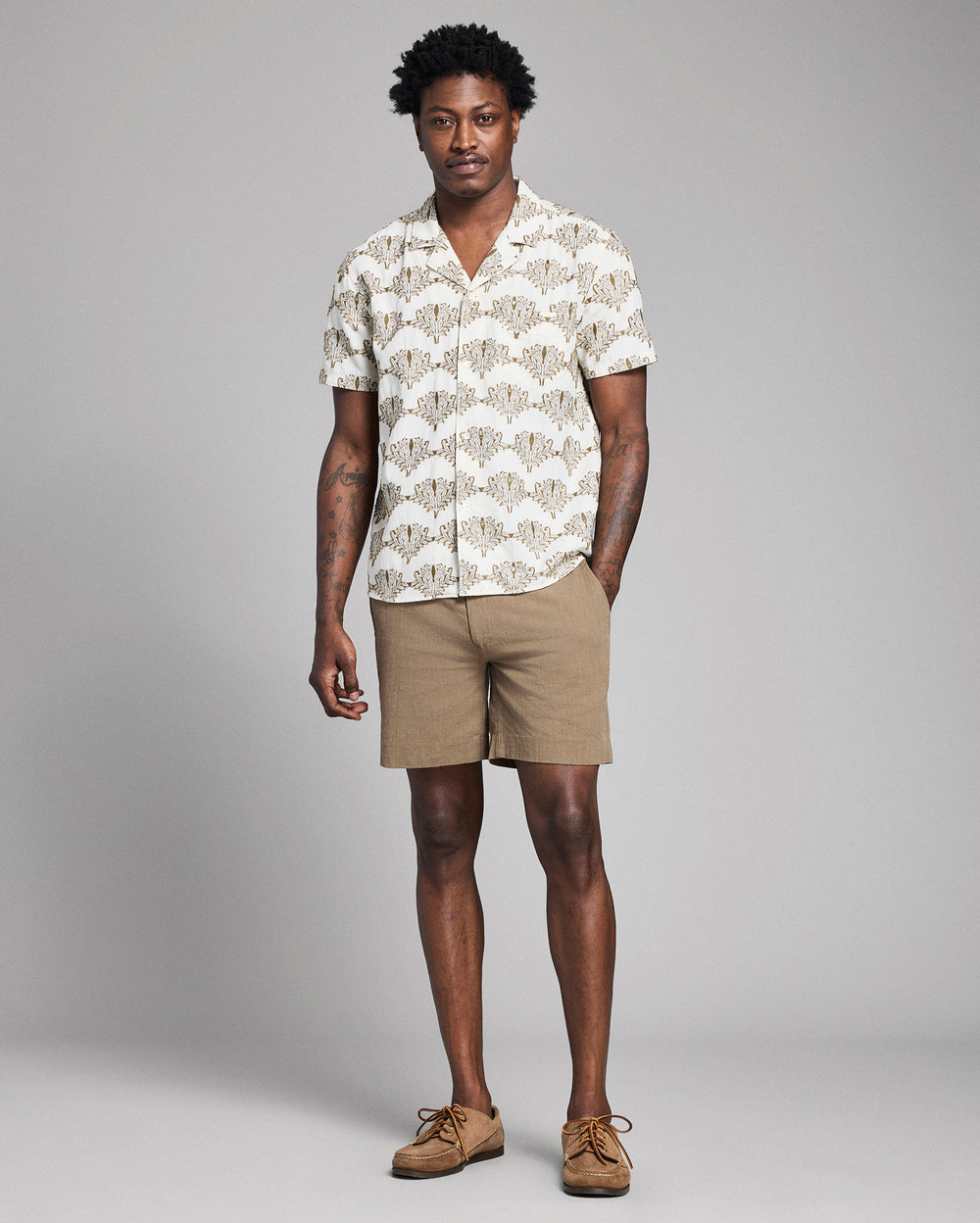 Man wearing a patterned shirt and beige shorts against a gray background