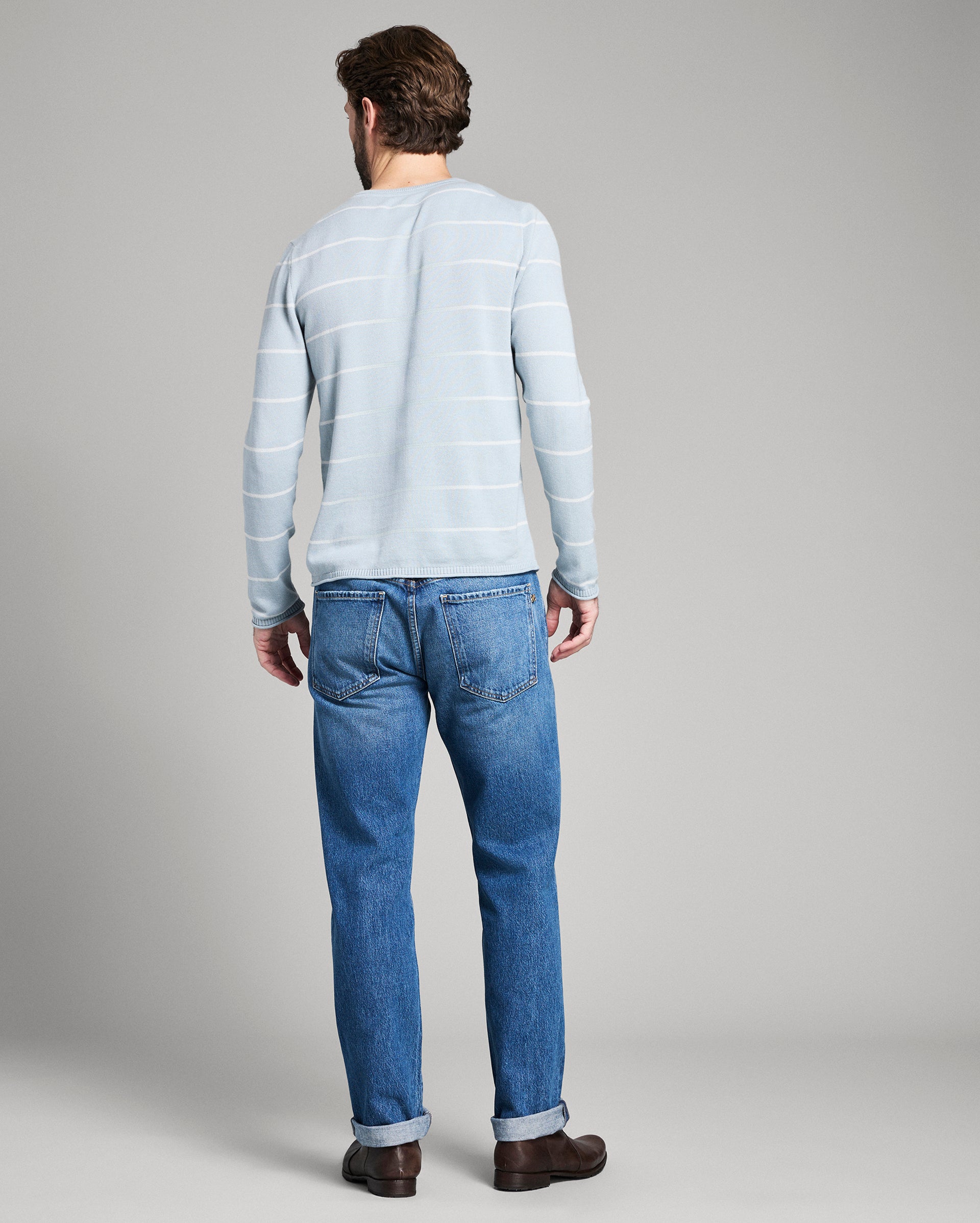 Man wearing a light blue striped sweater and blue jeans on a gray background