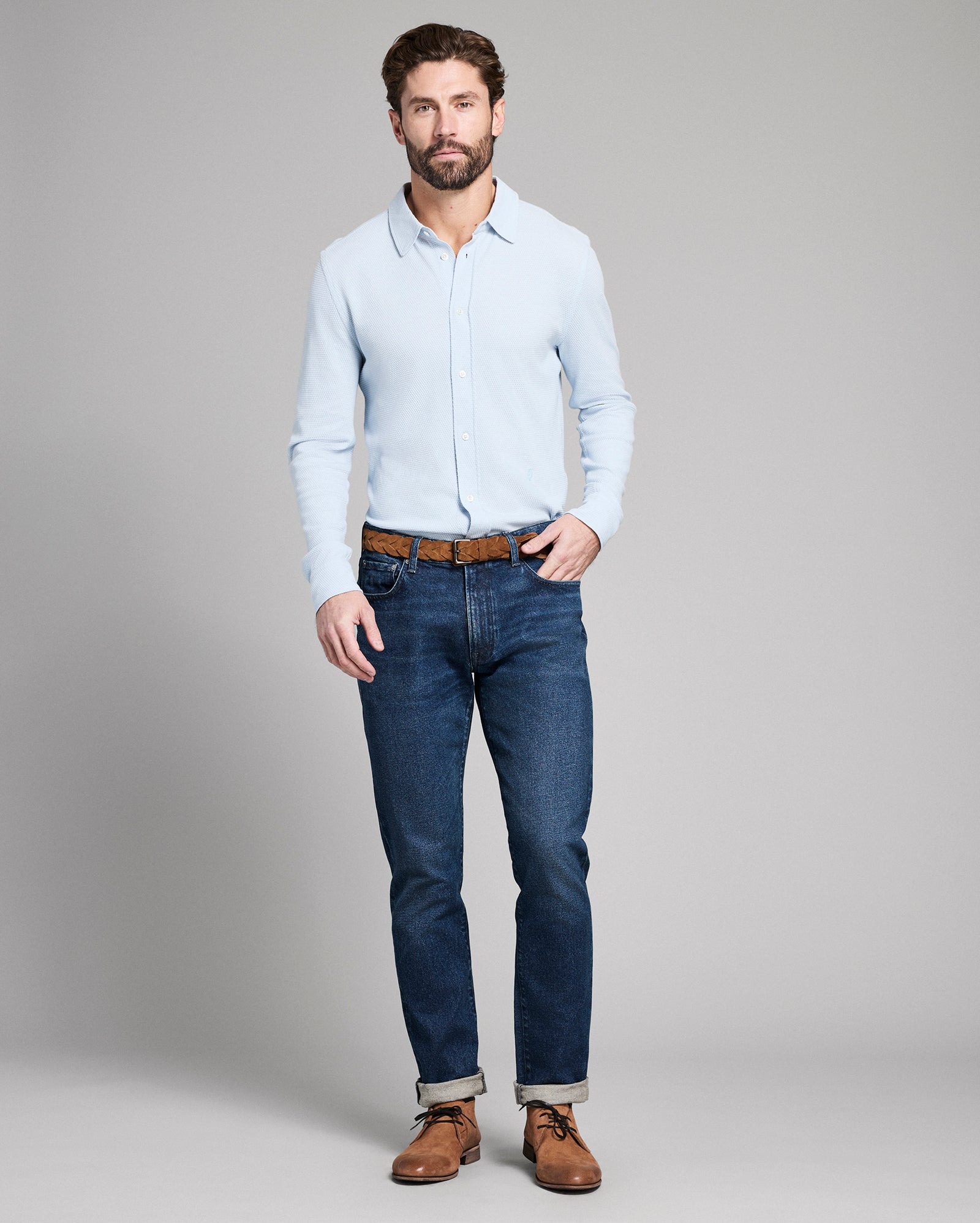 Man wearing a light blue shirt, dark blue jeans, and brown shoes on a gray background
