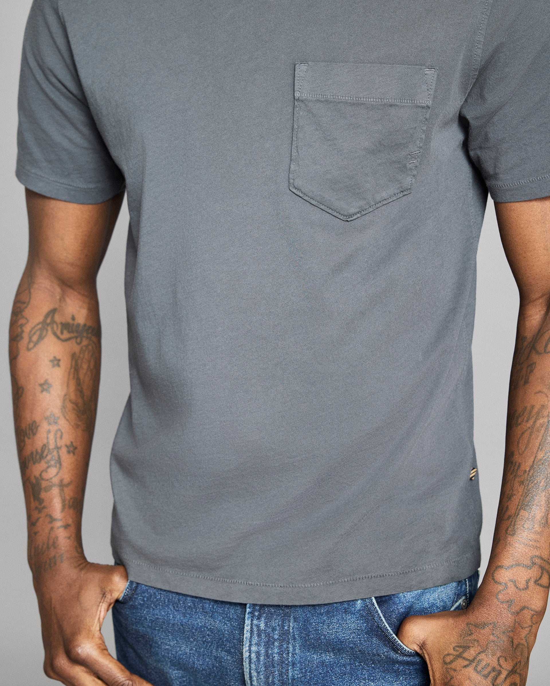 Gray t-shirt with a pocket worn by a person with tattoos on a plain background